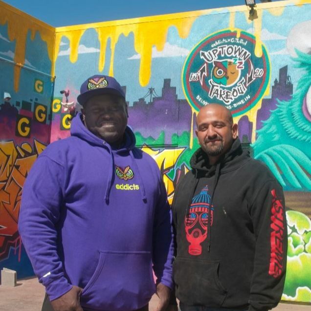 Photo of two men smiling in front of a mural that says, "Uptown Takeout."